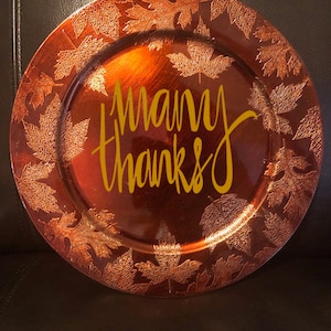 May include: A decorative, round plate with a metallic copper finish. The plate features a border of embossed leaves and the words "many thanks" in a yellow script. The plate is likely used for serving or display.