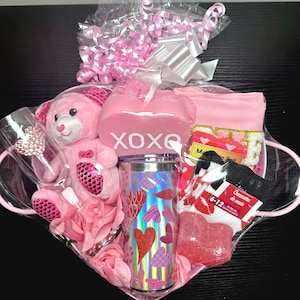 May include: A Valentine's Day gift basket filled with pink items. Includes a pink teddy bear, a tumbler with heart designs, a candle with "XOXO" text, socks, and a headband. The basket is wrapped in clear cellophane and tied with pink ribbons.