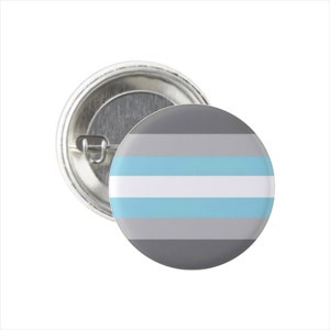 May include: A gray, white, and light blue button with a pin on the back. The button features the colors of the genderqueer pride flag.