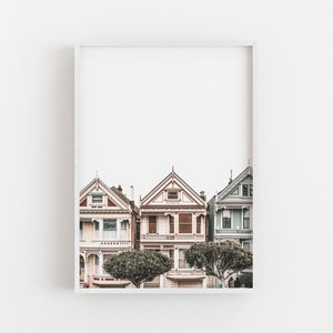 Puede incluir: Una impresi&oacute;n de arte mural enmarcada que muestra tres casas de estilo victoriano en San Francisco. Las casas est&aacute;n pintadas en tonos pastel suaves de rosa, crema y azul claro, con detalles arquitect&oacute;nicos intrincados y dos &aacute;rboles redondeados.