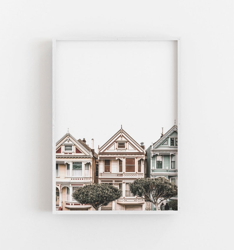 Puede incluir: Una impresi&oacute;n de arte mural enmarcada que muestra tres casas de estilo victoriano en San Francisco. Las casas est&aacute;n pintadas en tonos pastel suaves de rosa, crema y azul claro, con detalles arquitect&oacute;nicos intrincados y dos &aacute;rboles redondeados.