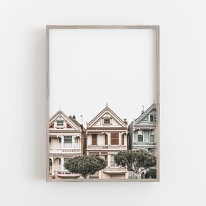 Puede incluir: Una impresi&oacute;n art&iacute;stica enmarcada que muestra tres casas de estilo victoriano en San Francisco. Las casas est&aacute;n pintadas en tonos rosa claro, crema y azul p&aacute;lido, con detalles arquitect&oacute;nicos intrincados, techos a dos aguas y dos &aacute;rboles.