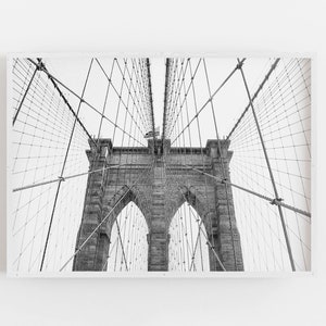 Puede incluir: Fotograf&iacute;a en blanco y negro del puente de Brooklyn, un puente colgante en Nueva York. La imagen captura las torres de piedra, los arcos y la intrincada red de cables del puente contra un cielo brillante. Una bandera americana es visible.