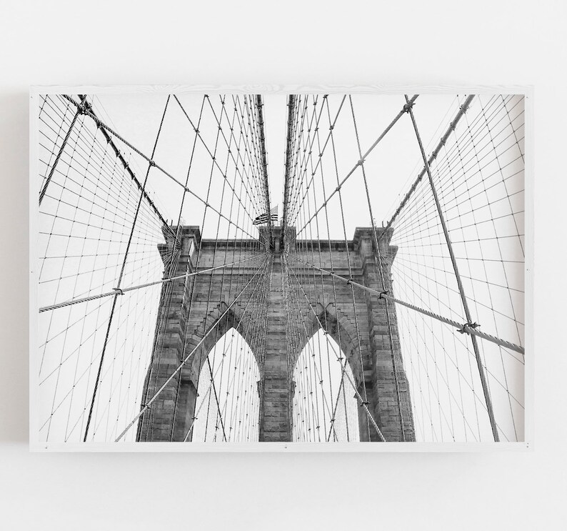 Puede incluir: Fotograf&iacute;a en blanco y negro del puente de Brooklyn, un puente colgante en Nueva York. La imagen captura las torres de piedra, los arcos y la intrincada red de cables del puente contra un cielo brillante. Una bandera americana es visible.