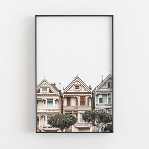 Puede incluir: Una impresi&oacute;n de arte mural enmarcada que muestra tres casas de estilo victoriano en San Francisco. Las casas est&aacute;n pintadas en tonos crema claro, blanco y azul p&aacute;lido con detalles arquitect&oacute;nicos intrincados, techos a dos aguas y dos &aacute;rboles verdes redondeados al frente.