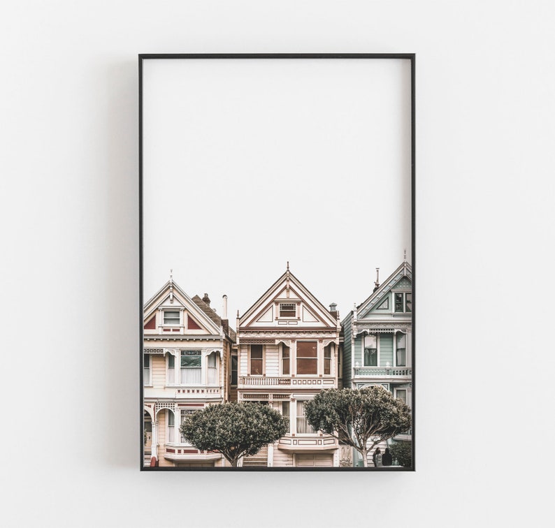 Puede incluir: Una impresi&oacute;n de arte mural enmarcada que muestra tres casas de estilo victoriano en San Francisco. Las casas est&aacute;n pintadas en tonos crema claro, blanco y azul p&aacute;lido con detalles arquitect&oacute;nicos intrincados, techos a dos aguas y dos &aacute;rboles verdes redondeados al frente.