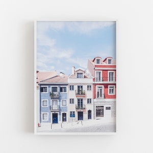 May include: A framed wall art print featuring a row of three multi-story buildings in blue, cream, and red. The blue building has patterned tile siding, the cream building has multiple balconies, and the red building has a stone-textured ground floor.