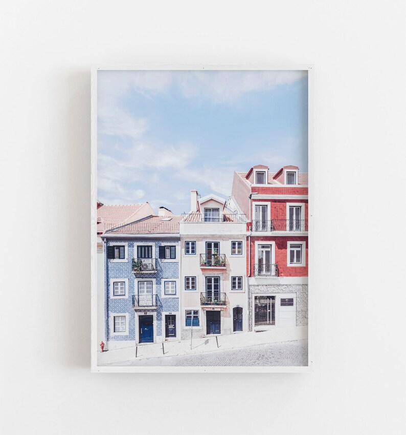 May include: A framed wall art print featuring a row of three multi-story buildings in blue, cream, and red. The blue building has patterned tile siding, the cream building has multiple balconies, and the red building has a stone-textured ground floor.