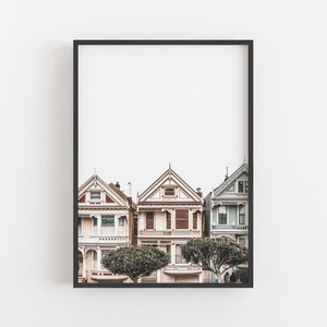 Puede incluir: Una impresi&oacute;n art&iacute;stica enmarcada que muestra tres casas de estilo victoriano en San Francisco. Las casas est&aacute;n pintadas en tonos rosa claro, crema y azul p&aacute;lido, con detalles arquitect&oacute;nicos intrincados, techos a dos aguas y dos &aacute;rboles verdes.