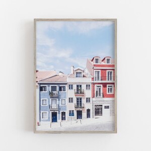 May include: A framed wall art print featuring a row of three multi-story buildings in blue, cream, and red. The blue building has patterned tiles, the cream building has balconies, and the red building has a stone base, all under a blue sky.