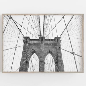 Puede incluir: Fotograf&iacute;a en blanco y negro del puente de Brooklyn, enmarcada en madera clara. La imagen captura las torres de piedra del puente, los cables de suspensi&oacute;n y la bandera estadounidense. La composici&oacute;n enfatiza los detalles arquitect&oacute;nicos del puente.