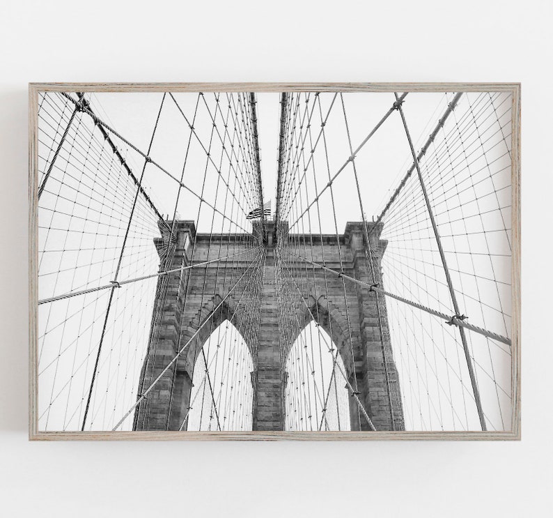 Puede incluir: Fotograf&iacute;a en blanco y negro del puente de Brooklyn, enmarcada en madera clara. La imagen captura las torres de piedra del puente, los cables de suspensi&oacute;n y la bandera estadounidense. La composici&oacute;n enfatiza los detalles arquitect&oacute;nicos del puente.