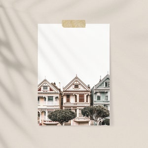 Puede incluir: Una l&aacute;mina de arte rectangular que muestra tres casas de estilo victoriano en tonos crema, marr&oacute;n claro y azul p&aacute;lido, con detalles arquitect&oacute;nicos intrincados y techos a dos aguas, fijada a una superficie neutra con cinta adhesiva beige.