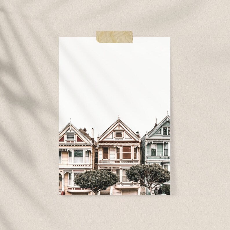 Puede incluir: Una l&aacute;mina de arte rectangular que muestra tres casas de estilo victoriano en tonos crema, marr&oacute;n claro y azul p&aacute;lido, con detalles arquitect&oacute;nicos intrincados y techos a dos aguas, fijada a una superficie neutra con cinta adhesiva beige.