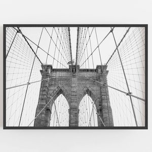 Puede incluir: Fotograf&iacute;a en blanco y negro del puente de Brooklyn, enmarcada en negro. Las torres de piedra y la intrincada red de cables del puente se ven contra un cielo brillante. Una bandera estadounidense ondea en la parte superior.
