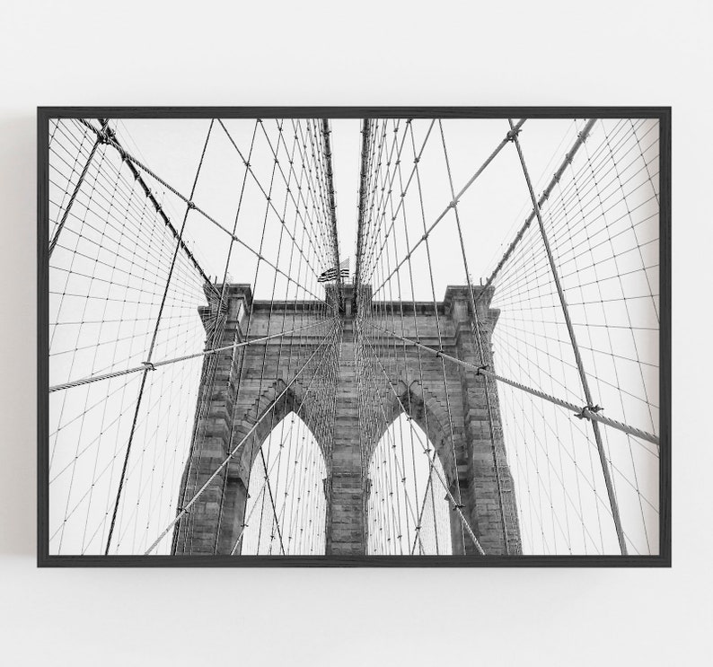 Puede incluir: Fotograf&iacute;a en blanco y negro del puente de Brooklyn, enmarcada en negro. Las torres de piedra y la intrincada red de cables del puente se ven contra un cielo brillante. Una bandera estadounidense ondea en la parte superior.