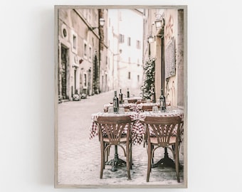 Puede incluir: Una fotografía vintage de un café en una calle estrecha y adoquinada de Italia. El café tiene manteles a cuadros rojos y blancos y sillas de madera. Hay botellas de vino en las mesas.