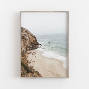 Może przedstawiać: Zdj?cie w ramie przedstawiaj?ce skalist? klif nad piaszczyst? pla??, na kt?rej fale rozbijaj? si? o brzeg. Niebo jest pochmurne, a woda ma jasnoniebieski kolor.