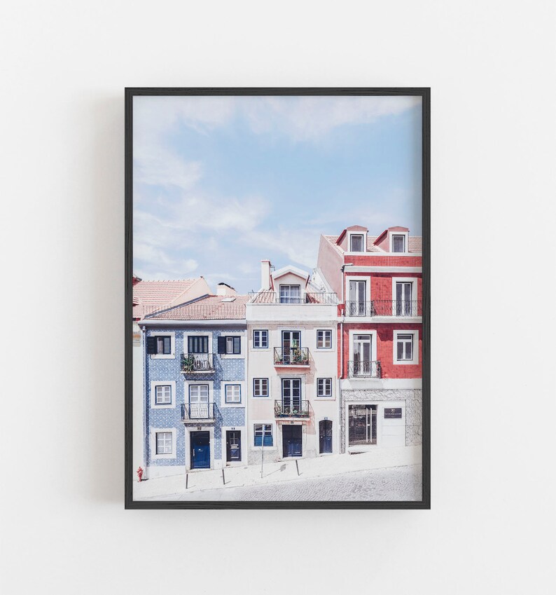 May include: A framed wall art print featuring a row of three multi-story buildings in blue, cream, and red. The blue building has patterned tile siding, the cream building has balconies, and the red building has a stone base and dark windows.