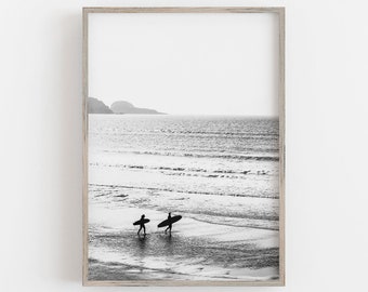 Puede incluir: Fotografía en blanco y negro de dos personas caminando por una playa con tablas de surf. Las personas están silueteados contra el agua y el cielo. La playa es arenosa y el agua está tranquila.