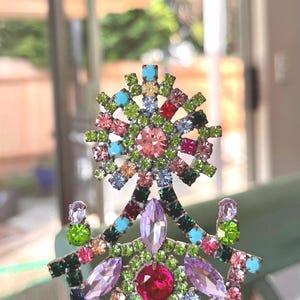 May include: A decorative brooch featuring a starburst top section and a lower structure adorned with an array of multi-colored rhinestones. The piece includes round, marquise, and square-cut crystals in shades of lime green, pink, red, blue, and purple set in a metallic frame.