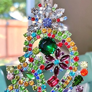 May include: A decorative brooch featuring a dense arrangement of multi-colored rhinestones in shades of red, green, blue, yellow, purple, and clear. The piece includes a large central green stone and various floral-shaped metal settings.