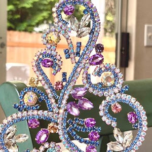 May include: A decorative ornament featuring an intricate, swirling metal frame encrusted with numerous multi-colored rhinestones. The design includes various shades of blue, purple, pink, and clear crystals arranged in a complex, branching pattern.