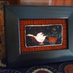 Puede incluir: Obra de arte enmarcada que representa un ganso en vuelo sobre un carro rojo, sobre un cielo nocturno negro con estrellas. La obra de arte está enmarcada en un marco rectangular negro, con un fondo rojo texturizado. A la izquierda se ve otra foto enmarcada.