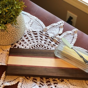 May include: A rectangular wooden cutting board with a light brown and purple stripe across the center. A clear glass butter dish with a silver butter knife is on the board.
