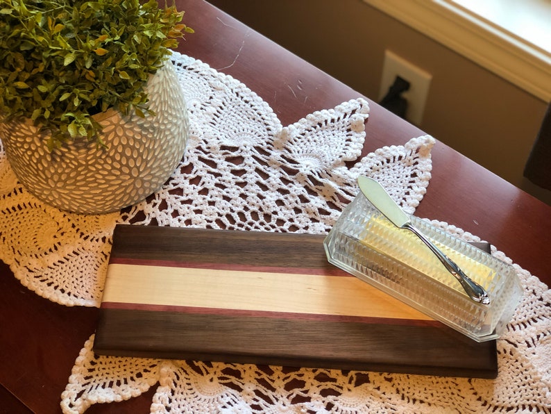 May include: A rectangular wooden cutting board with a light brown and purple stripe across the center. A clear glass butter dish with a silver butter knife is on the board.