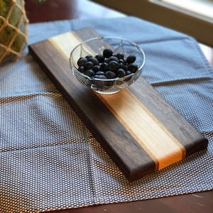 May include: A rectangular wooden serving board with a light wood center stripe and dark wood sides. A small glass bowl filled with blueberries sits on the board.