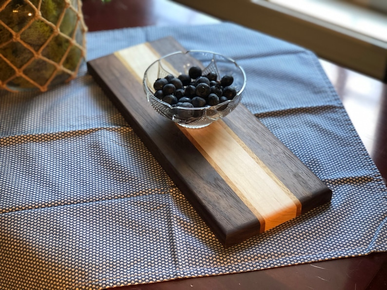 May include: A rectangular wooden serving board with a light wood center stripe and dark wood sides. A small glass bowl filled with blueberries sits on the board.