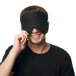 Puede incluir: Una persona con una máscara de dormir negra sobre los ojos, sonriendo y mirando a la cámara. La máscara está hecha de un tejido suave y cómodo y tiene un diseño sencillo.