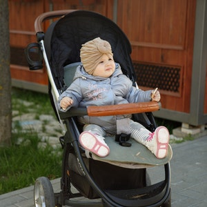 May include: A black stroller with a brown leather-look handle and bumper bar, carrying a child wearing a beige turban-style hat and a metallic silver puffer jacket with the text Be a star B.S. printed on the chest, paired with striped leggings.
