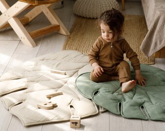 May include: A child sits on a leaf-shaped play mat. The mat is light green and beige, with a quilted design. Wooden blocks with numbers and the word "October" are nearby. The child wears a brown hooded jumpsuit.