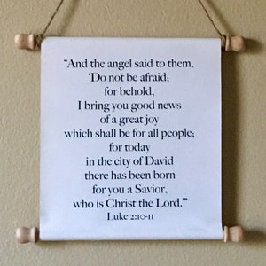 May include: A white scroll with black text, hanging from a string. The text is a passage from the Bible, Luke 2:10-11, about the birth of Jesus Christ. The scroll is a religious decoration for a home or office.