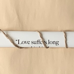 May include: A white scroll with the text "Love suffers long" is rolled up and tied with twine. The scroll is attached to two wooden dowels with rounded ends.