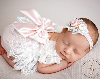 May include: A pale pink romper featuring a large satin bow on the back and intricate white lace trim along the edges. The outfit is paired with a matching white elastic headband adorned with small pink and white fabric flowers and lace details.