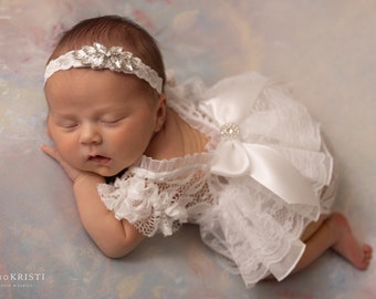 May include: A sleeping newborn baby wearing a white lace dress with a large white satin bow and a silver headband with rhinestones.