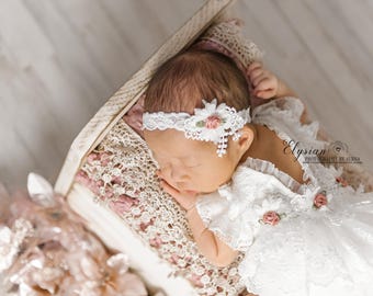 May include: A newborn baby girl sleeping in a white lace dress with a white lace headband with a pink flower. The baby is lying on a pink and white lace blanket in a white wooden crib.