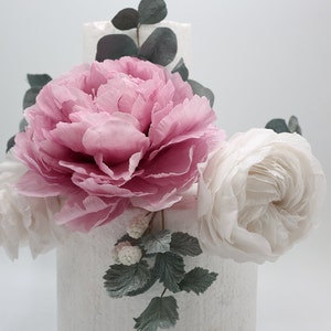 May include: A two-tiered white cake decorated with pink and white paper flowers. The cake is adorned with greenery and small white berries.