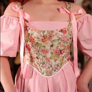 May include: A pink dress with a floral bodice featuring fairies, flowers, and butterflies. The dress has off-the-shoulder sleeves and pink ribbon straps. The skirt is pleated, and the overall style is reminiscent of a fairy tale.