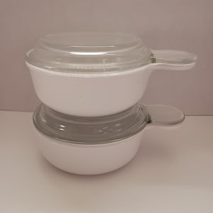May include: Two white, handled bowls with clear glass lids are stacked. The bowls have a rounded shape and a single handle on each side. The lids are clear and fit snugly on top of the bowls. The bowls are likely for cooking or serving.