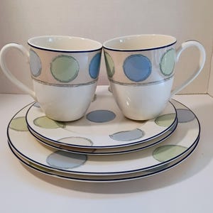 May include: Two white ceramic mugs and two plates with a polka dot design. The mugs and plates have a blue, green, and gray color scheme. The mugs have a handle and a blue trim. The plates have a blue trim.