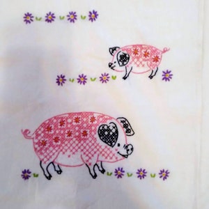 May include: White fabric featuring two embroidered pink pigs with black details and heart-shaped eyes. The pigs are decorated with cross-stitch patterns and surrounded by purple flower embroidery.