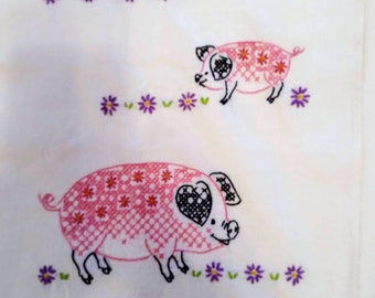 May include: White fabric featuring two embroidered pink pigs with black details and heart-shaped eyes. The pigs are decorated with cross-stitch patterns and surrounded by purple flower embroidery.