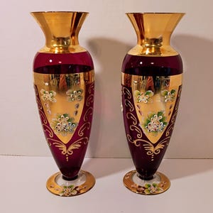 May include: Two decorative glass vases. The vases have a deep red body with gold accents on the rim, base, and decorative panels. The panels feature hand-painted floral designs in white and green.