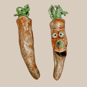 Puede incluir: Dos figuritas de zanahoria de cerámica con caras pintadas. Una zanahoria tiene una parte superior verde frondosa y una cara pintada con ojos grandes y una amplia sonrisa. La otra zanahoria tiene una parte superior verde frondosa y un cuerpo naranja liso.