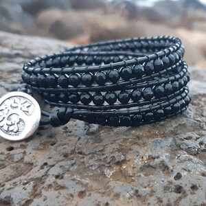 May include: Black beaded wrap bracelet with a silver tree of life charm. The bracelet is made of multiple strands of black beads and is tied with a black leather cord.