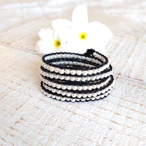 May include: A black and white beaded wrap bracelet with white pearls. The bracelet is made of multiple strands of black cord and white pearls.
